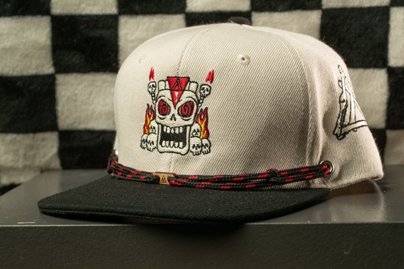 A white baseball cap with a skull design and a black and red braided strap is displayed against a black and white checkered background.
