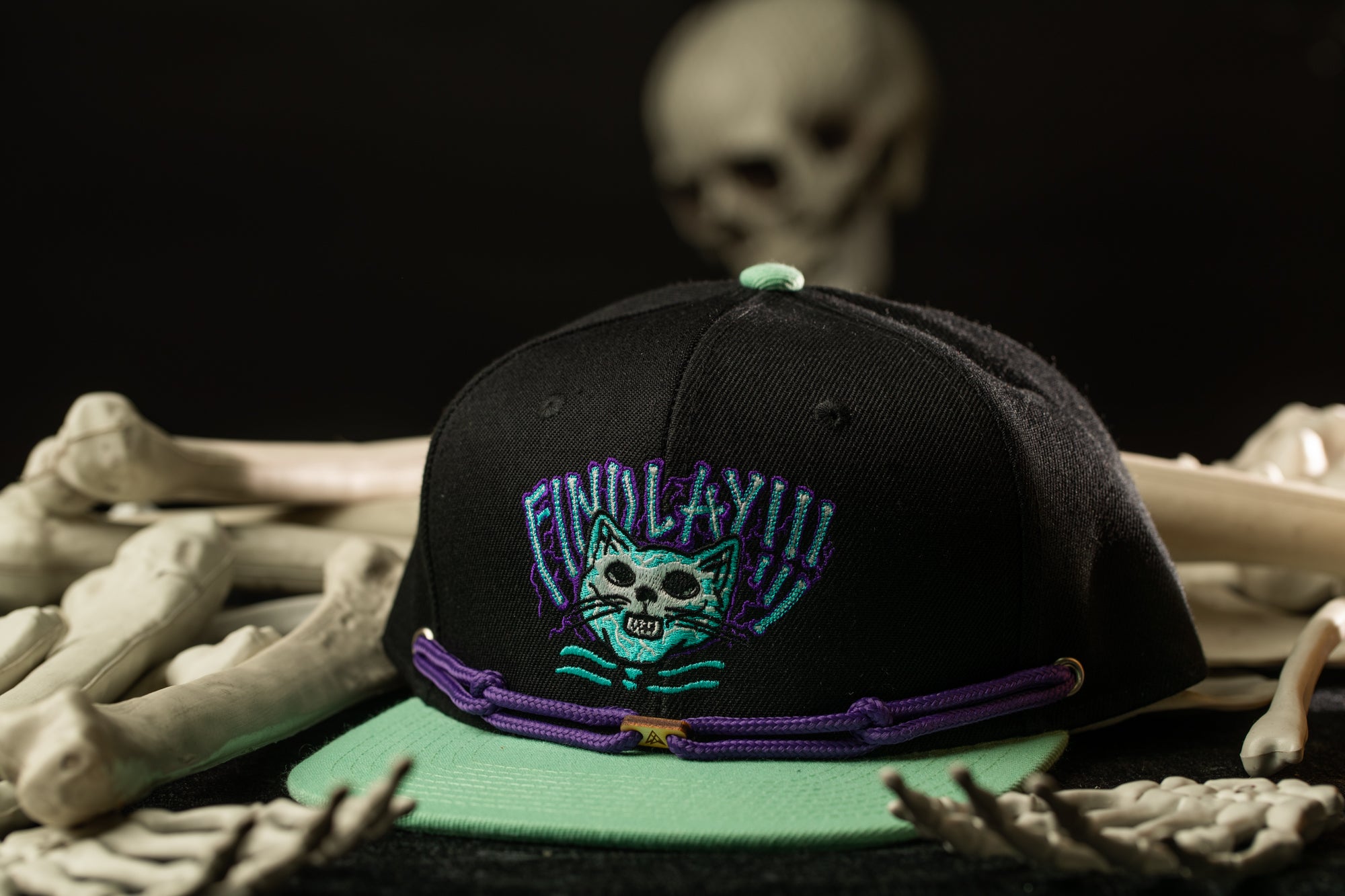 A black baseball cap with a purple and teal design, featuring a skull and crossbones motif, is placed on a surface with a pile of bones and a blurred skull in the background.