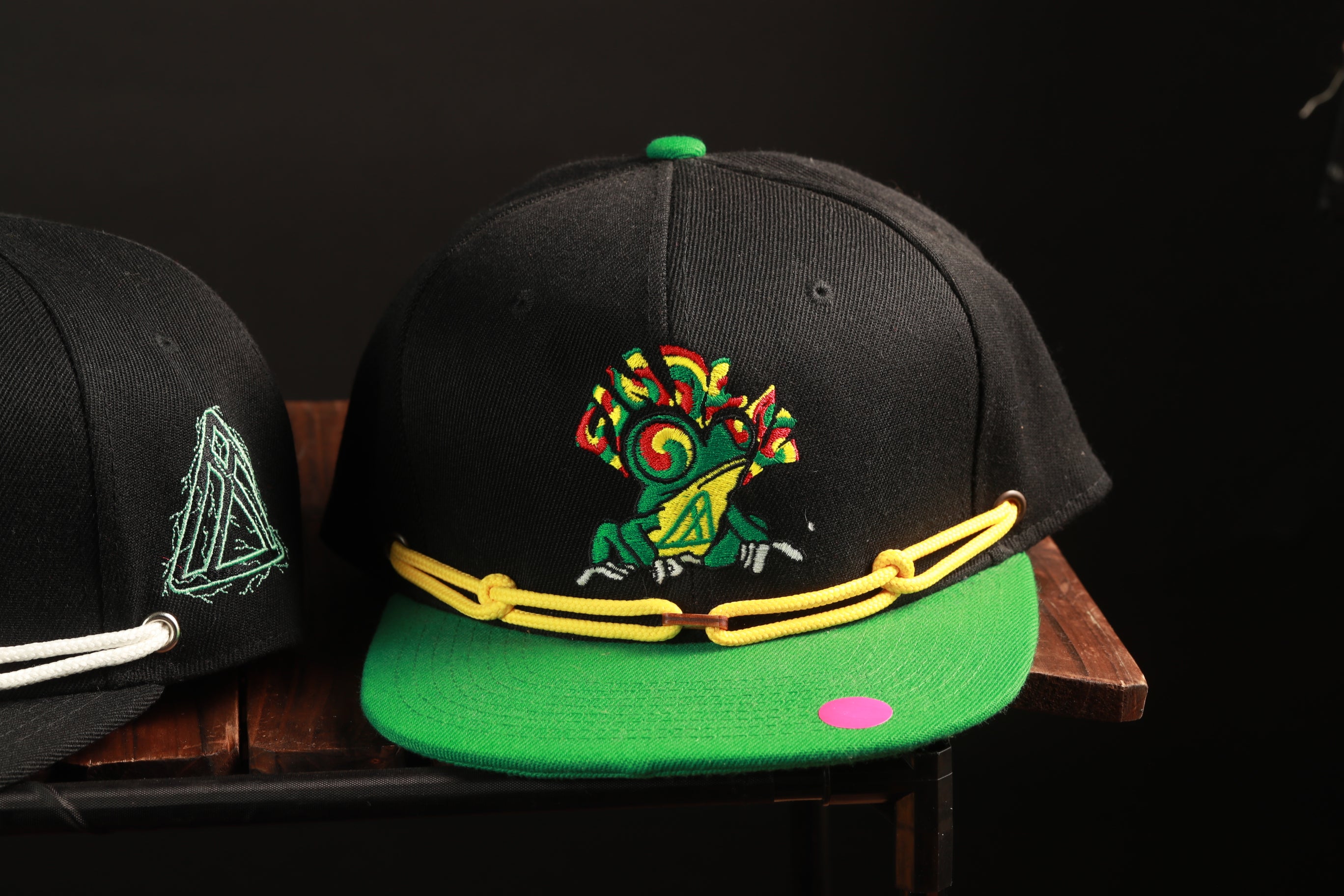 A black baseball cap with a green brim and a colorful frog design on the front, sitting on a wooden surface.