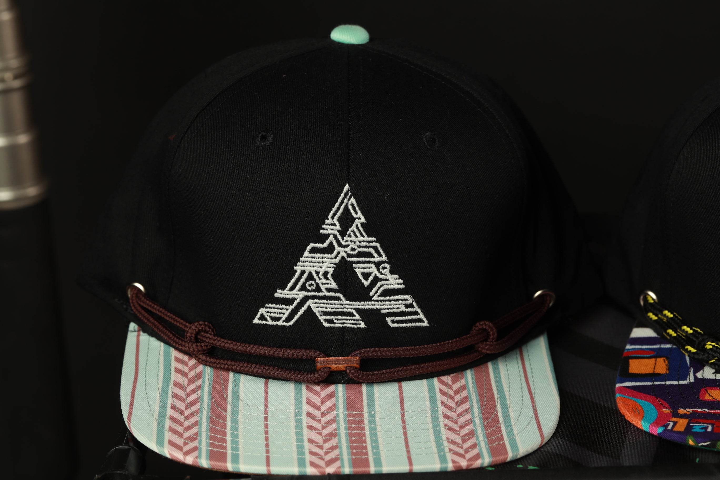 A black baseball cap with a white graphic design on the front, featuring a stylized letter 'A' and a striped pattern on the brim.