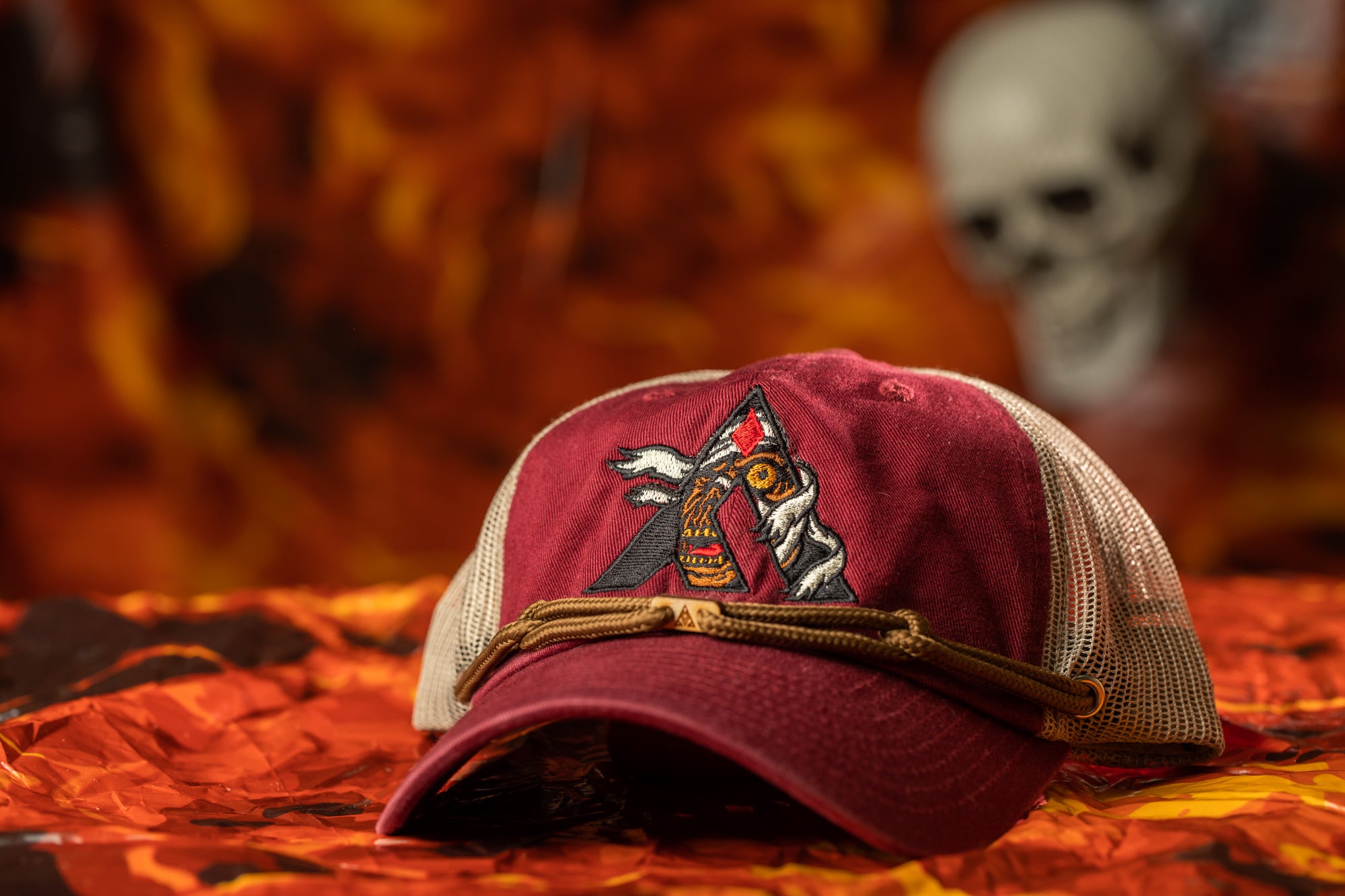 A maroon baseball cap with a graphic design on the front, placed on a surface with orange and brown leaves. In the background, there is a blurred image of a skull.