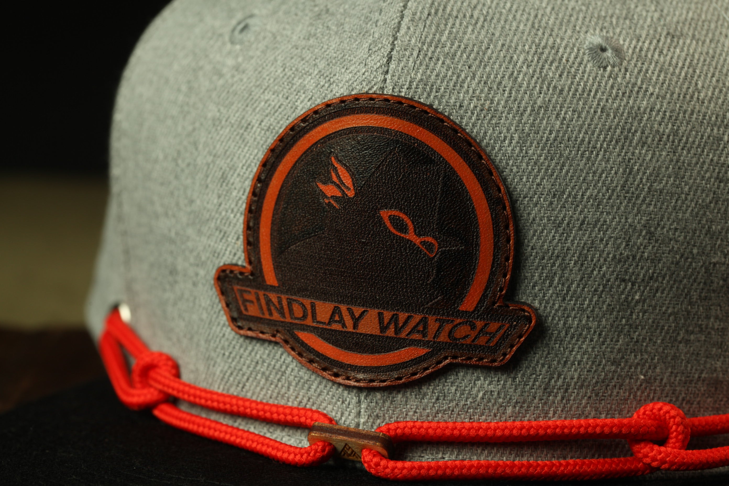 A gray baseball cap with a red chain and a circular patch on the front displaying the text "FINDLAY WATCH".