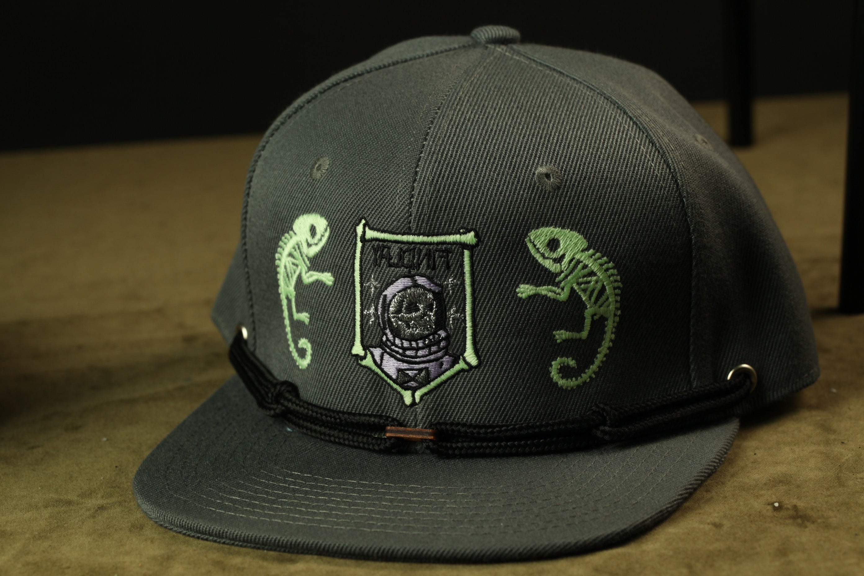 A gray baseball cap with a black band and a patch featuring a skull and chameleon designs.