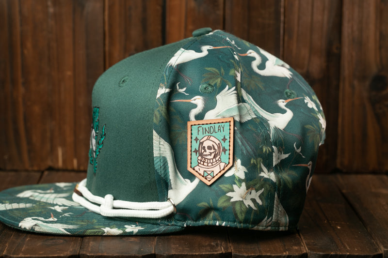 A green baseball cap with a floral pattern and a skull patch on the front, placed on a wooden surface.