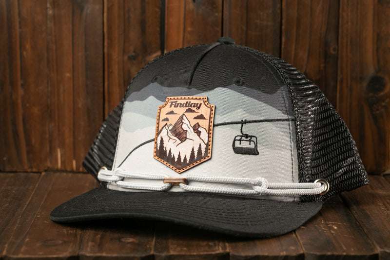 A black and gray baseball cap with a mountain and ski lift design, placed on a wooden surface.