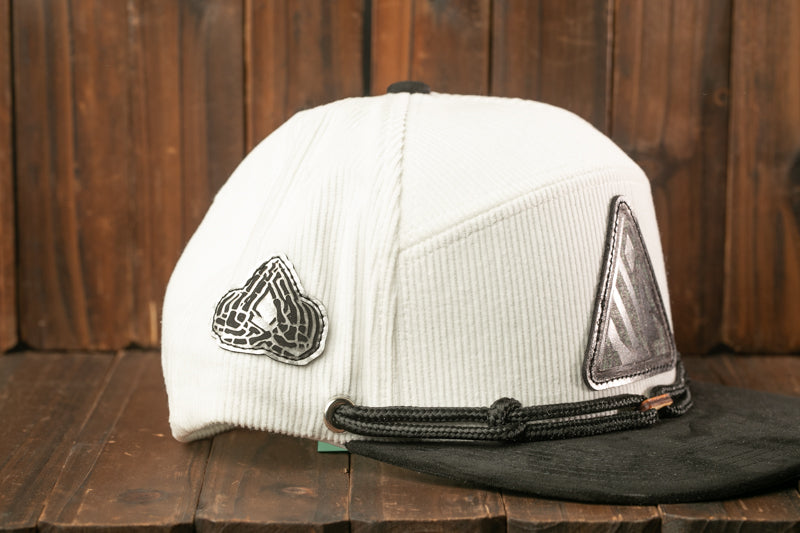 A white baseball cap with a black brim and a black and white geometric patch on the front, placed on a wooden surface.