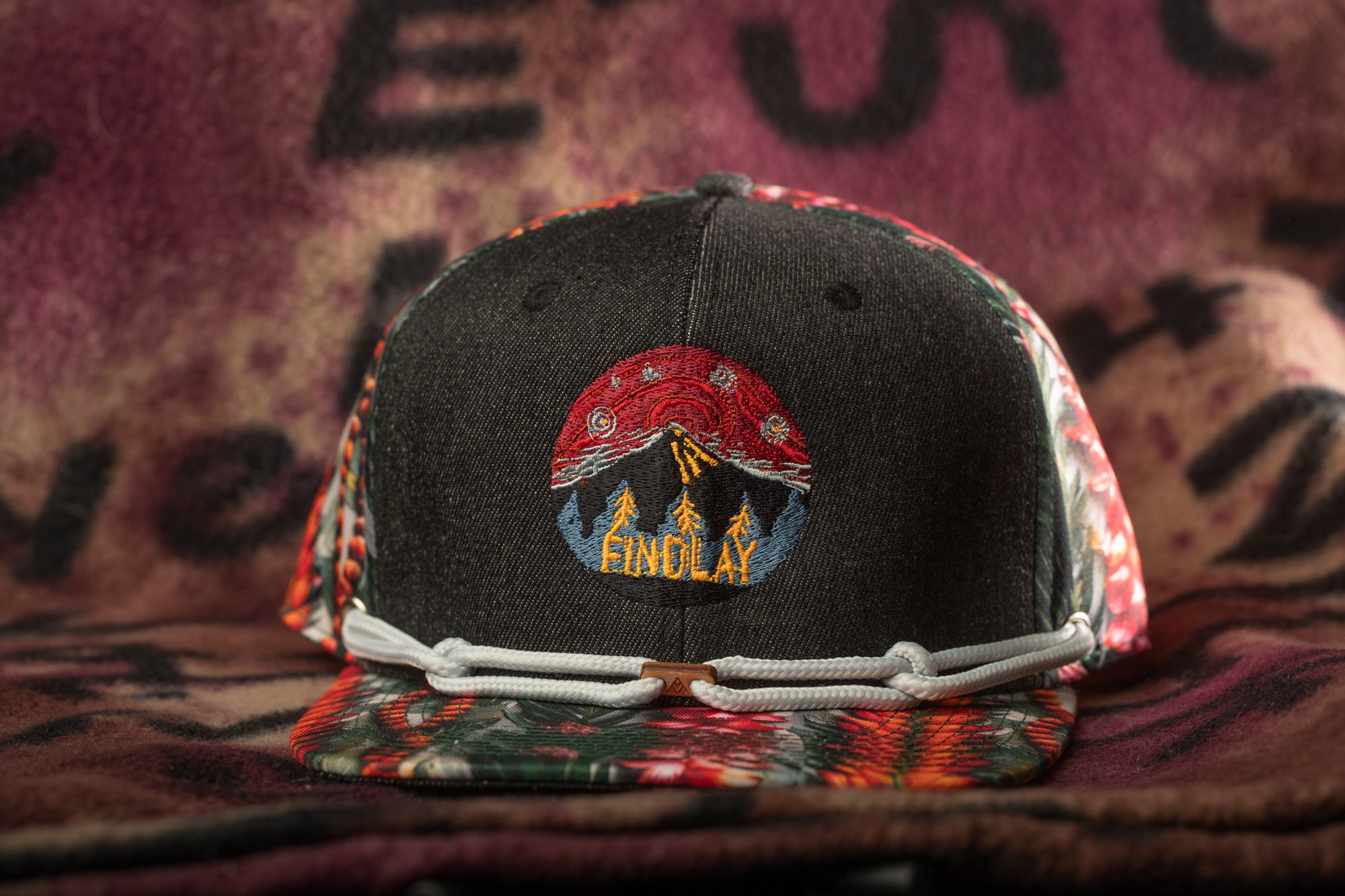 A black baseball cap with a colorful, embroidered design on the front, featuring a mountain landscape and the word "FINOLAY".