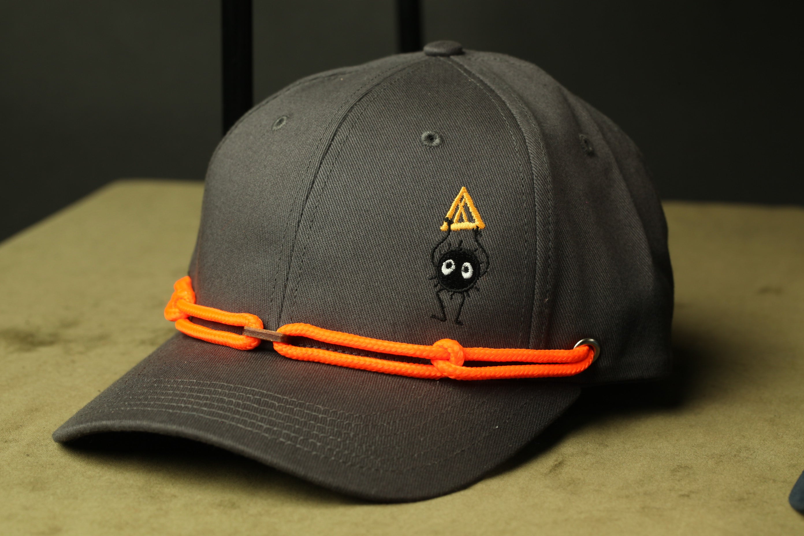 A black baseball cap with an orange string and a small cartoon spider design on the front.