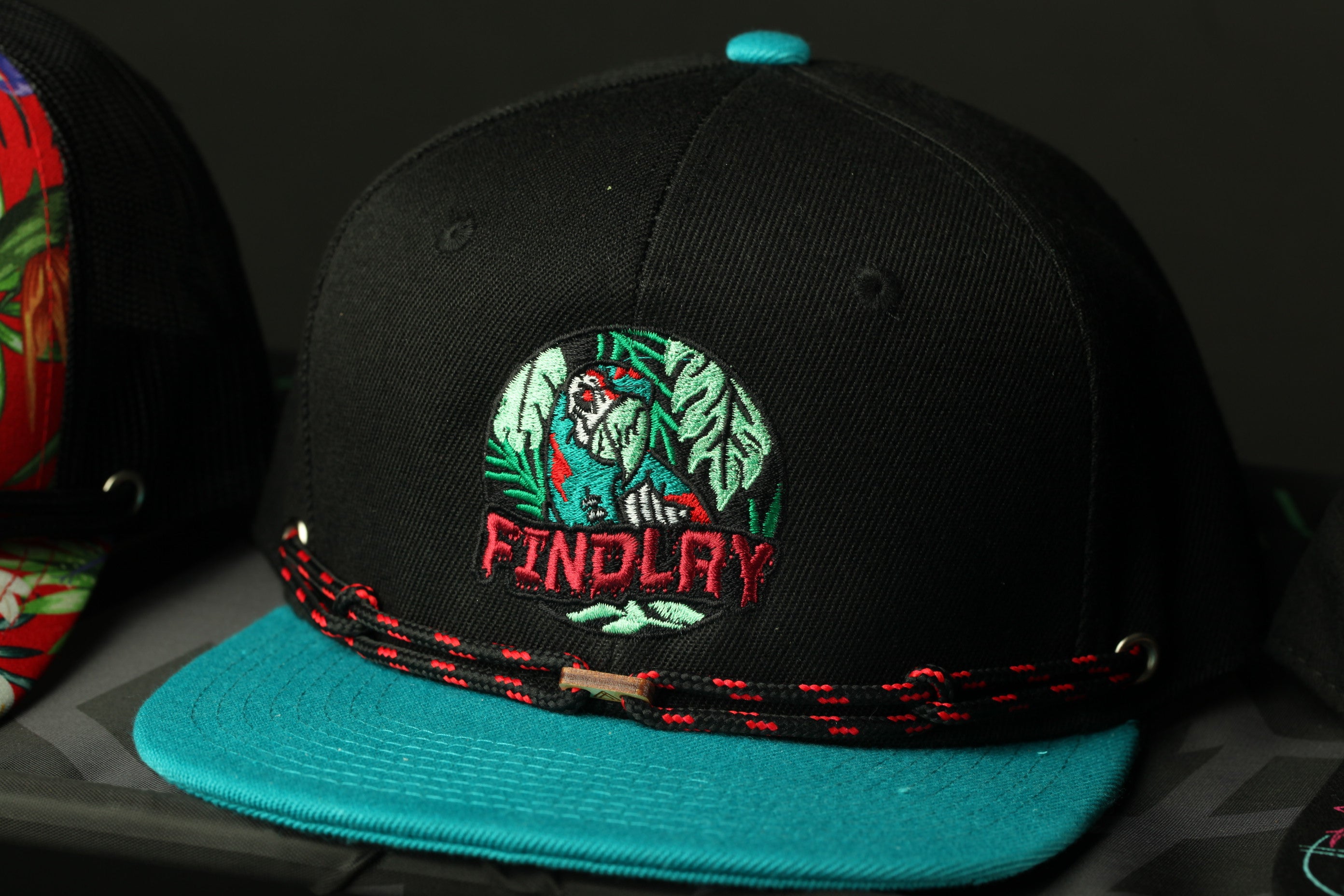 A black baseball cap with a teal brim and a red and green embroidered logo that says "FINDLAY".