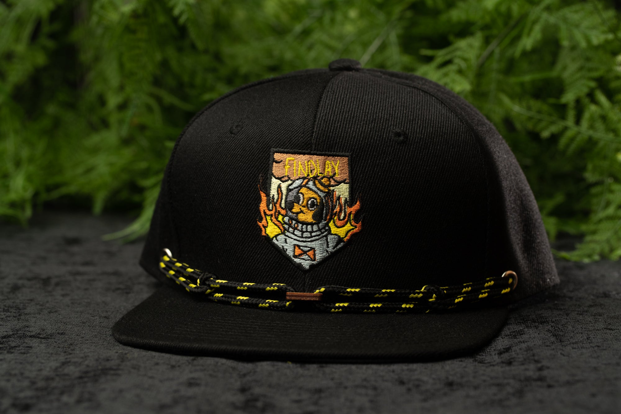A black baseball cap with a yellow and orange design on the front, featuring a shield-shaped emblem with flames and a skull.