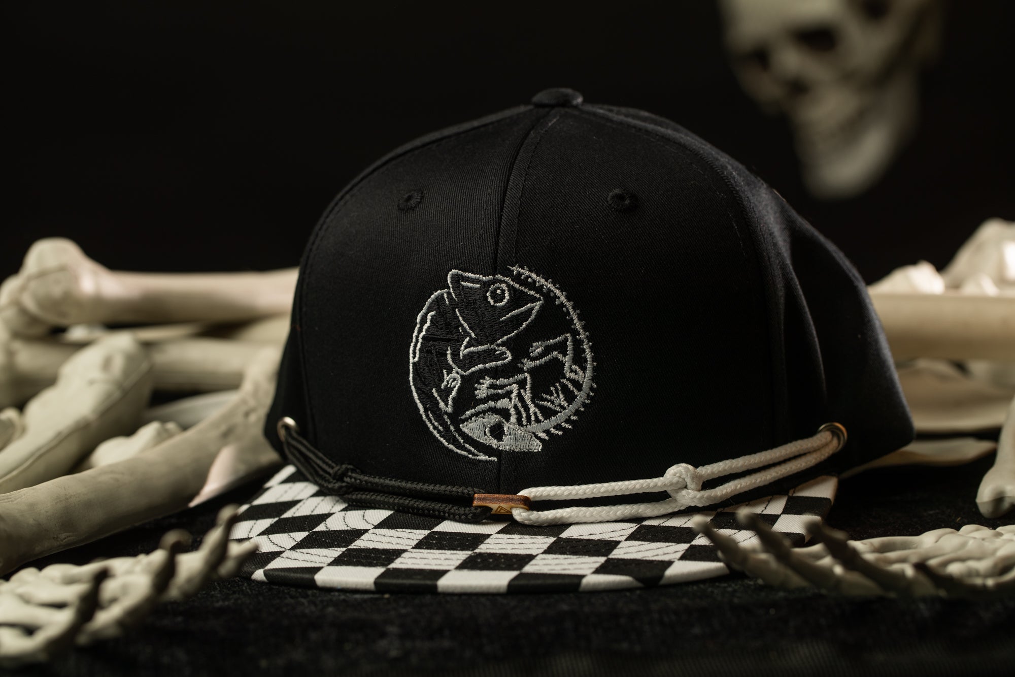A black baseball cap with a white embroidered design sits on a checkered patterned surface, surrounded by scattered bones.