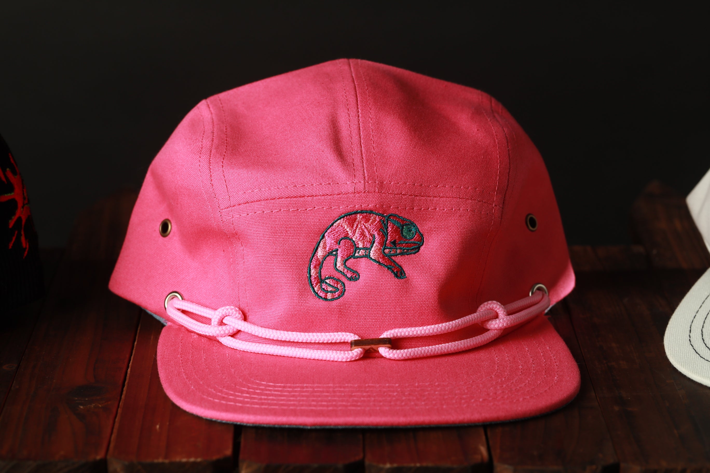 A pink baseball cap with a chameleon embroidered on it, sitting on a wooden surface.