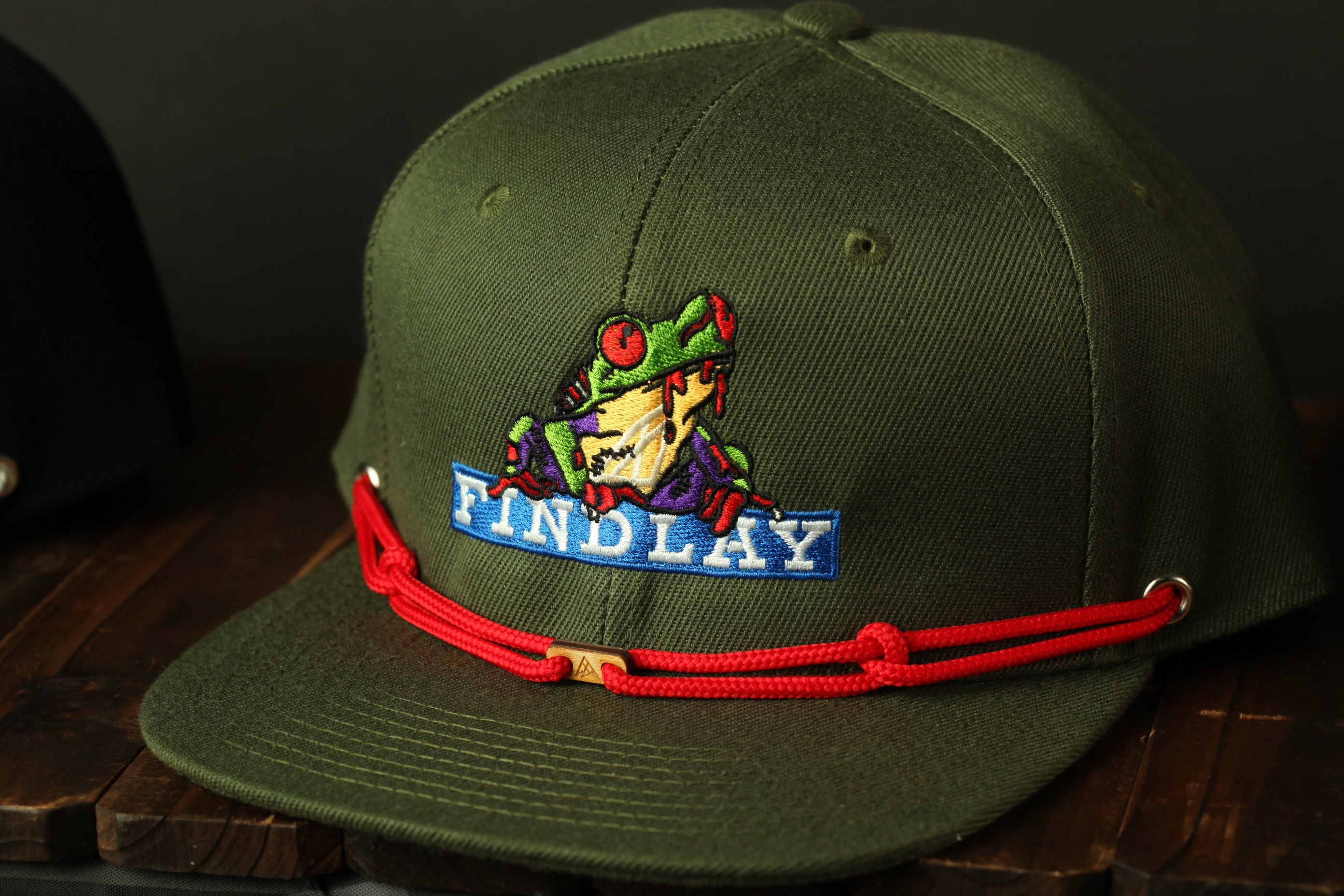 A green baseball cap with a red string and a frog embroidered on it, sitting on a wooden surface.