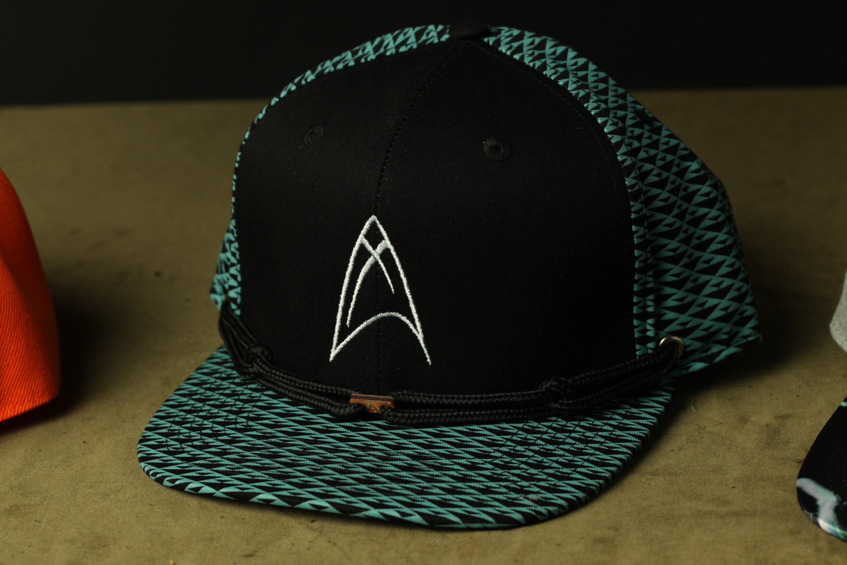 A black baseball cap with a teal and white patterned design, featuring a white logo on the front.