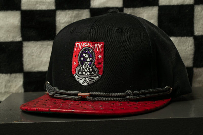 A black baseball cap with a red brim and a red and white logo on the front, placed on a black surface against a black and white checkered background.