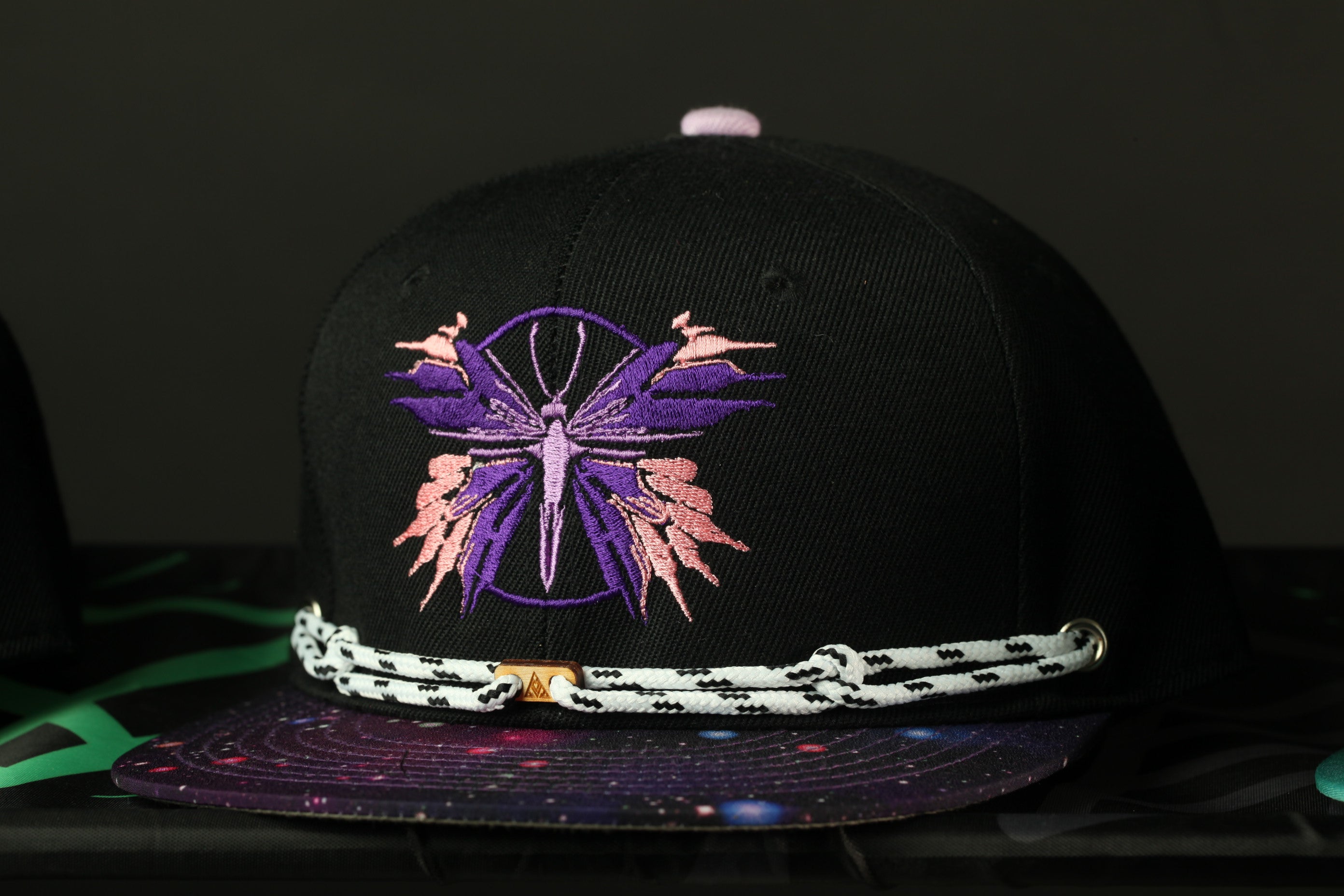 A black baseball cap with a purple and pink butterfly embroidered on it, sitting on a black surface with a colorful galaxy pattern.