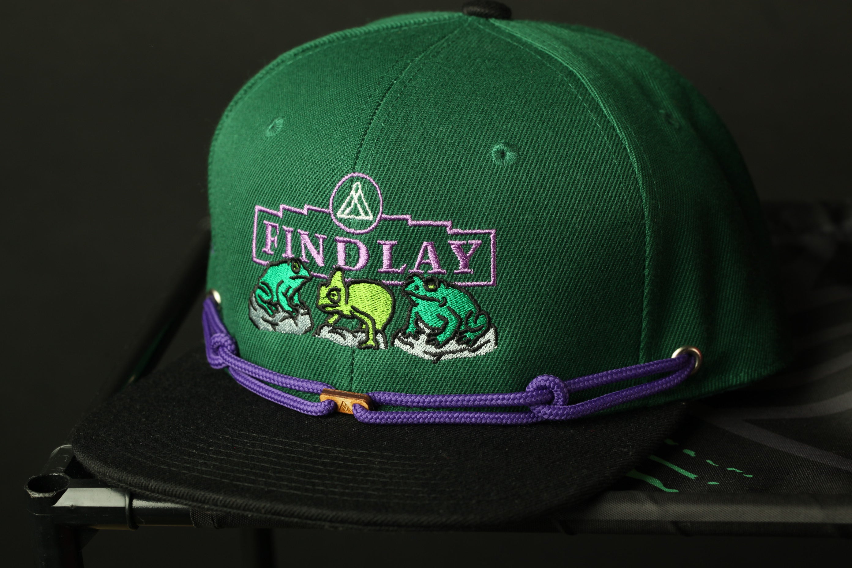 A green baseball cap with a purple band and a logo on it, sitting on a black surface.