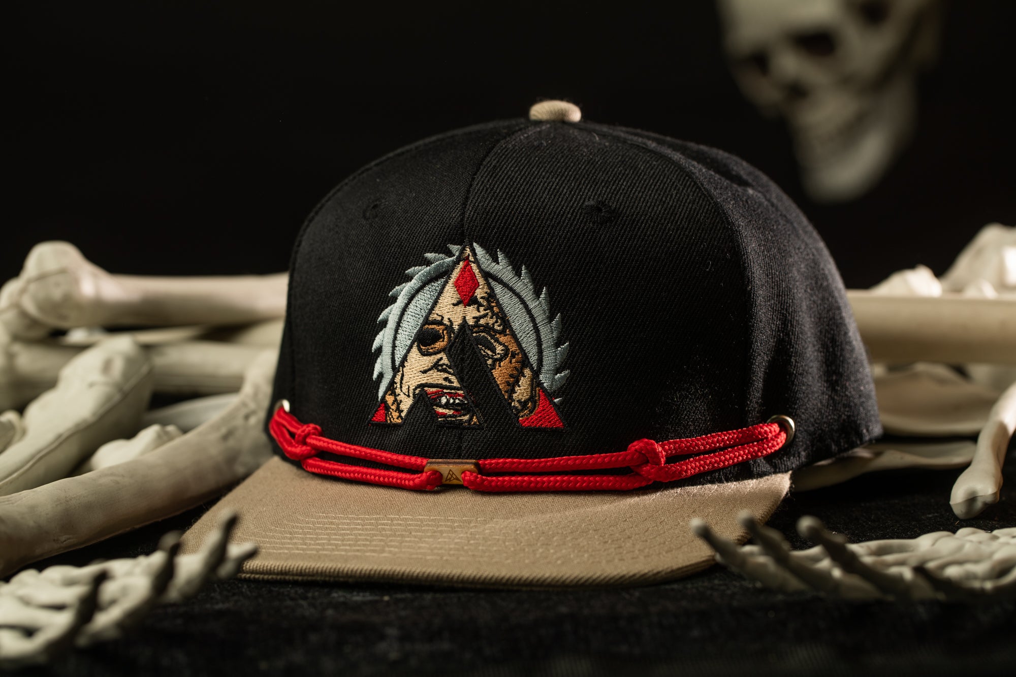 A black baseball cap with a red band and an embroidered design on the front, placed on a surface with bones and a skull in the background.