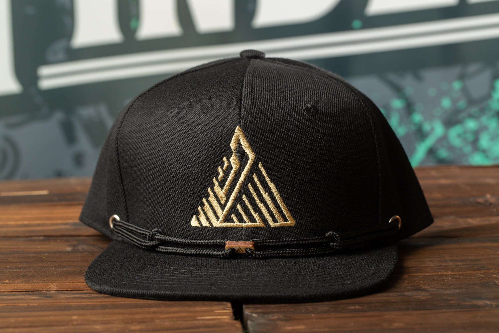 A black baseball cap with a gold geometric logo on the front, placed on a wooden surface.