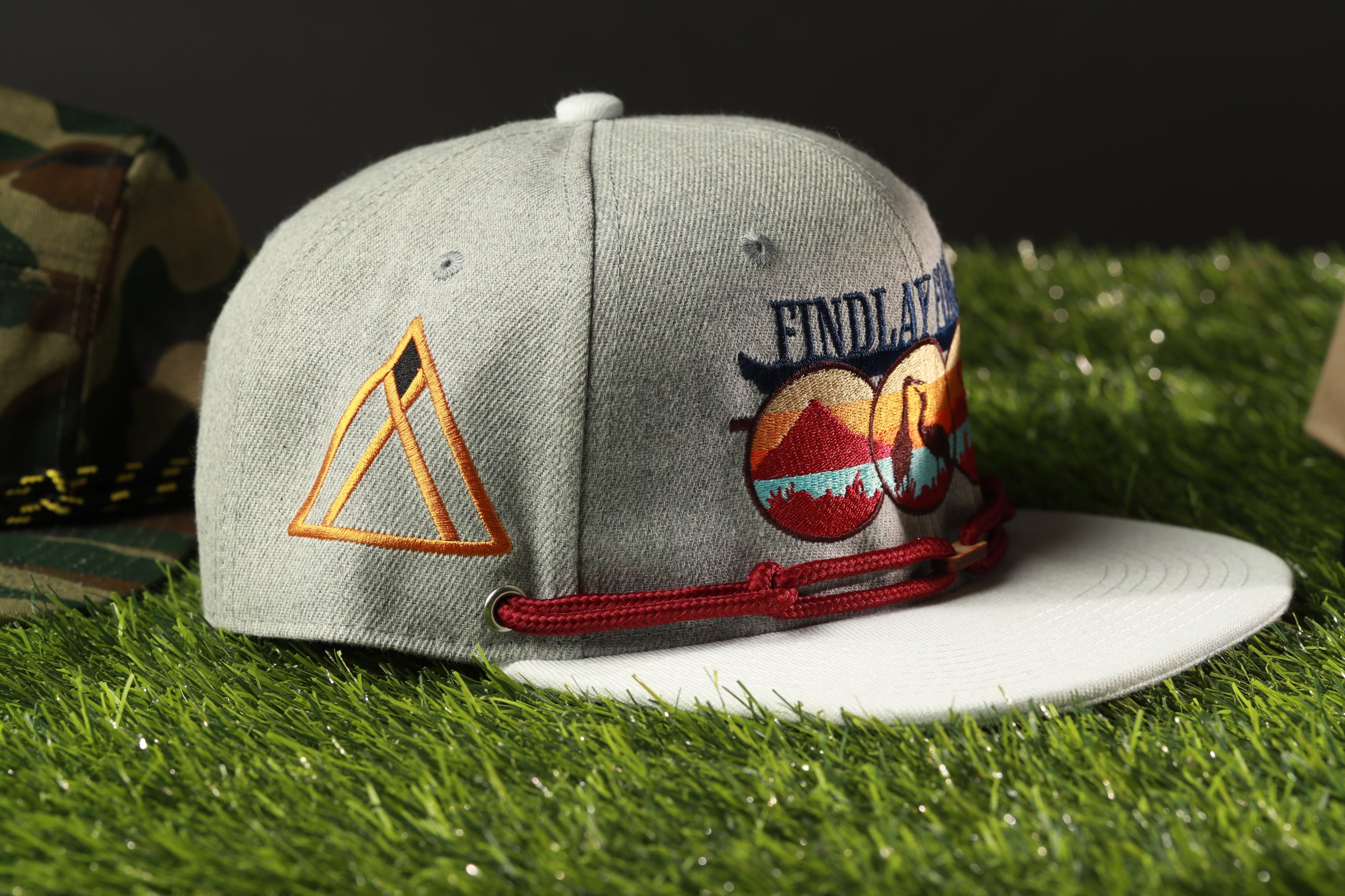 A gray baseball cap with a red string and a triangle design on the side, placed on a grassy surface.