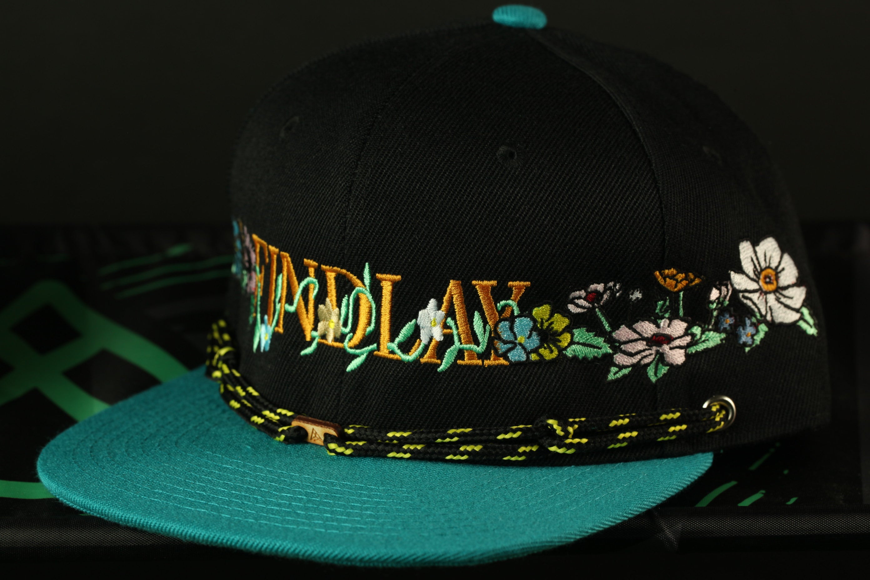 A black baseball cap with a teal brim and floral embroidered text on the front.