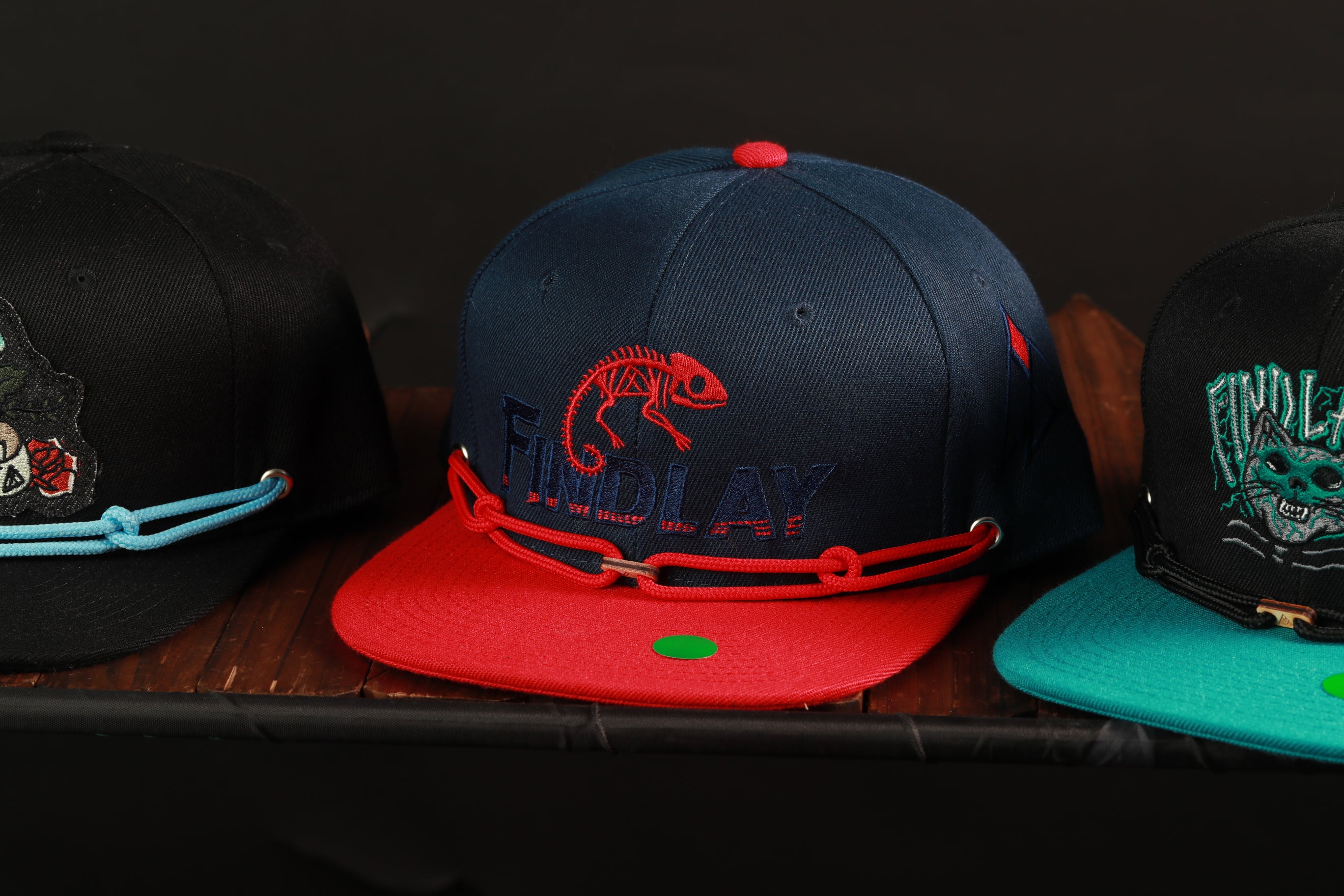 Three baseball caps are displayed on a wooden surface, each with a unique design and color.