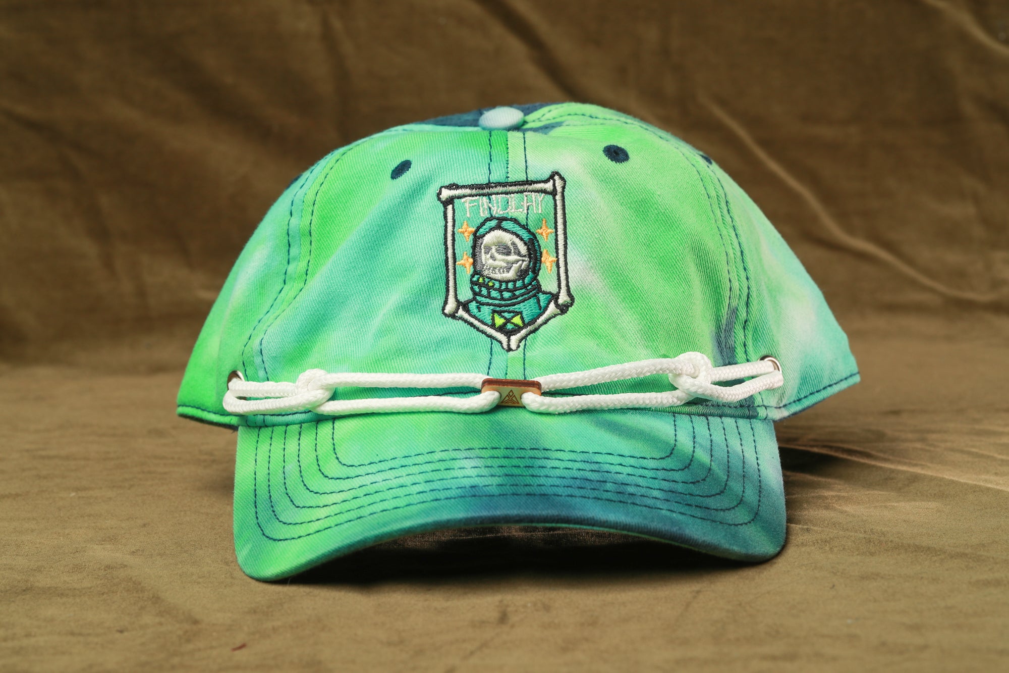 A tie-dye baseball cap with a skull and crossbones design on the front, featuring a white rope strap.
