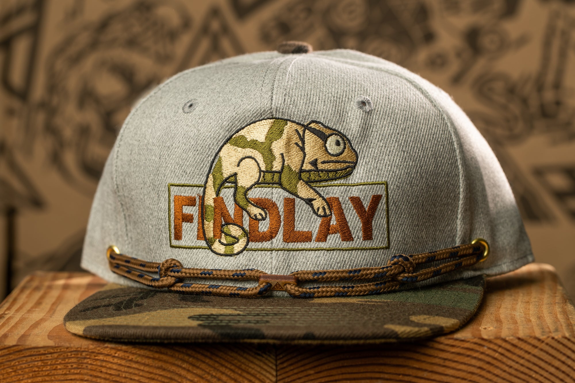 A gray baseball cap with a camouflage pattern and a chameleon design, along with the word "FINDlay" printed on it.