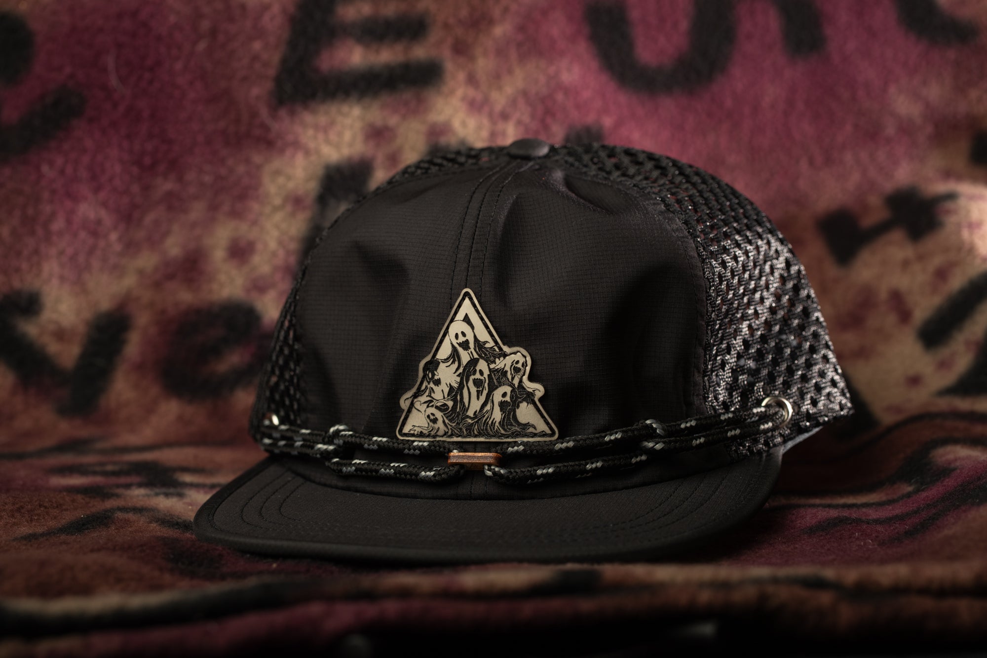 A black baseball cap with a mesh back and a triangular logo on the front, placed on a patterned fabric background.